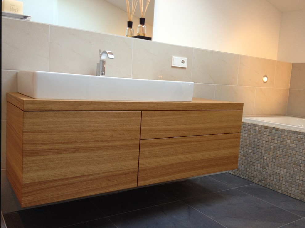 Cette image montre une salle de bain minimaliste en bois foncé de taille moyenne avec un placard à porte plane, une baignoire en alcôve, WC suspendus, un carrelage beige, un carrelage en pâte de verre, un mur blanc, parquet en bambou, une vasque et un plan de toilette en bois.