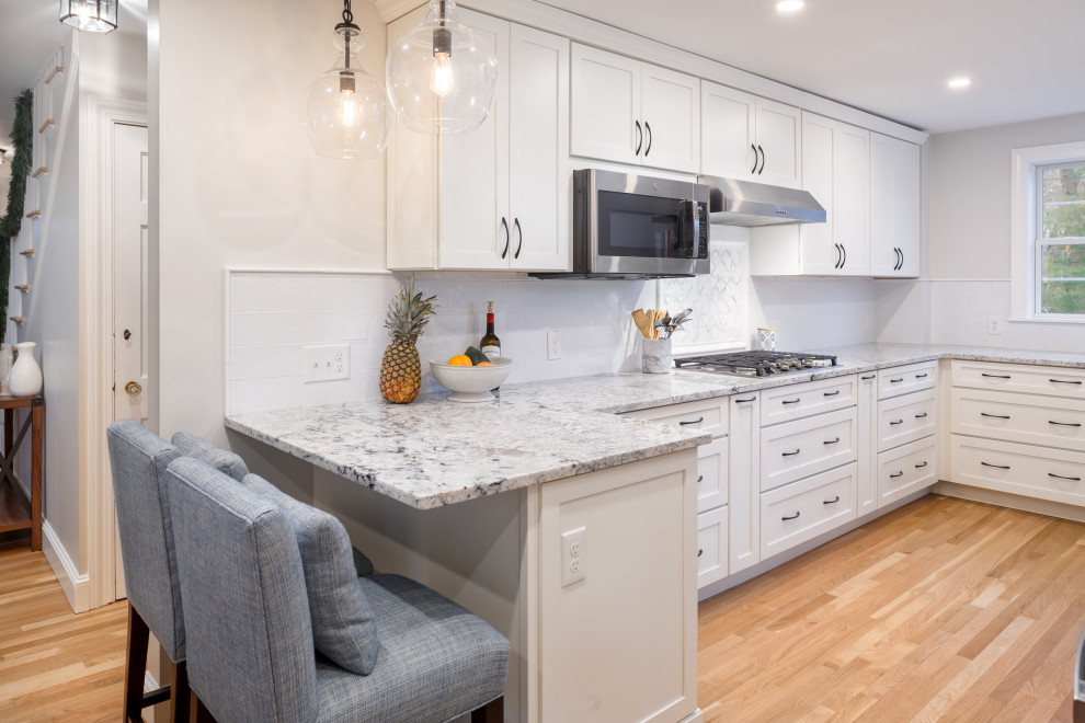 Inspiration for a mid-sized transitional u-shaped medium tone wood floor eat-in kitchen remodel in Boston with a farmhouse sink, shaker cabinets, white cabinets, granite countertops, white backsplash, subway tile backsplash, stainless steel appliances, a peninsula and multicolored countertops