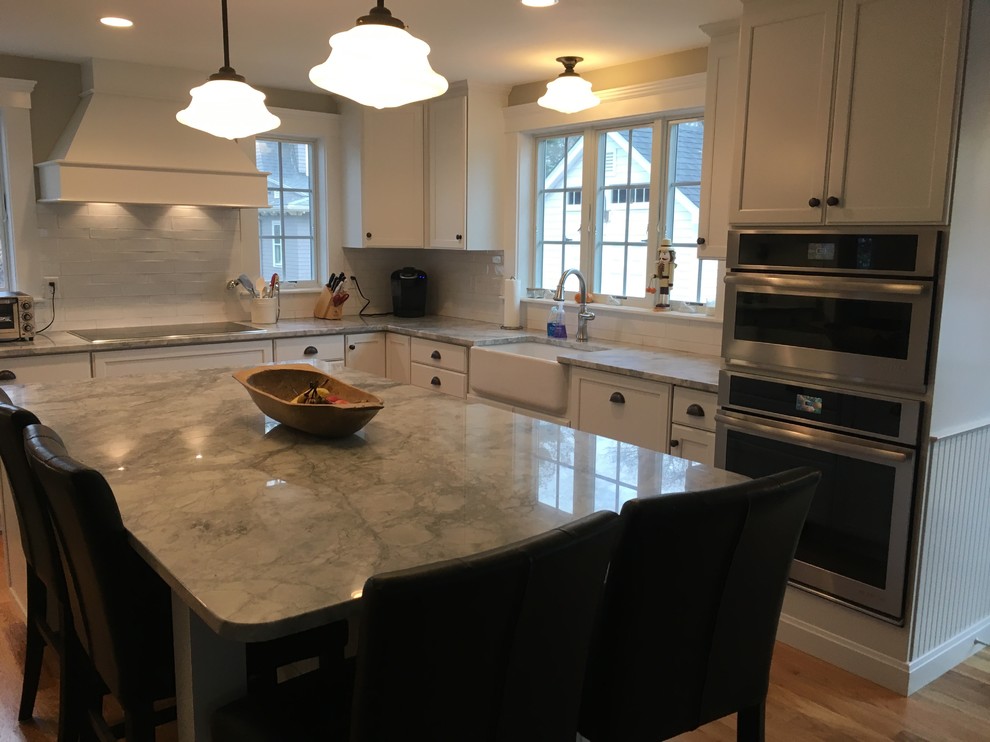 Example of a transitional l-shaped medium tone wood floor eat-in kitchen design in Boston with a farmhouse sink, shaker cabinets, white cabinets, white backsplash, porcelain backsplash, stainless steel appliances and an island