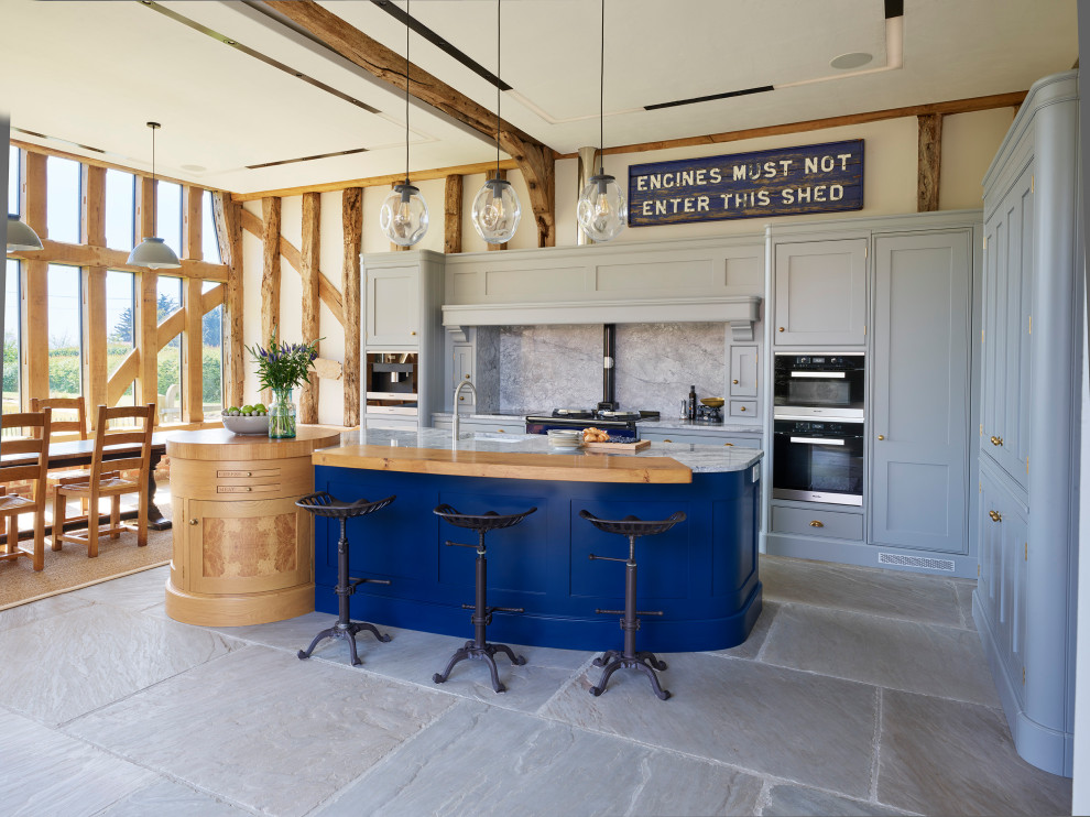This is an example of a rural l-shaped kitchen/diner in Essex with a belfast sink, shaker cabinets, grey cabinets, granite worktops, grey splashback, granite splashback, black appliances, an island, grey floors and grey worktops.