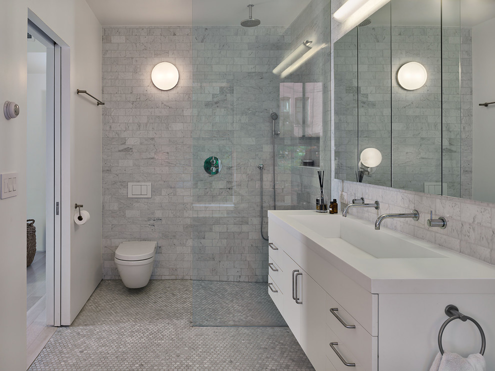 This is an example of a mid-sized contemporary master bathroom in New York with flat-panel cabinets, white cabinets, an open shower, a wall-mount toilet, gray tile, grey walls, a trough sink, grey floor, an open shower and white benchtops.