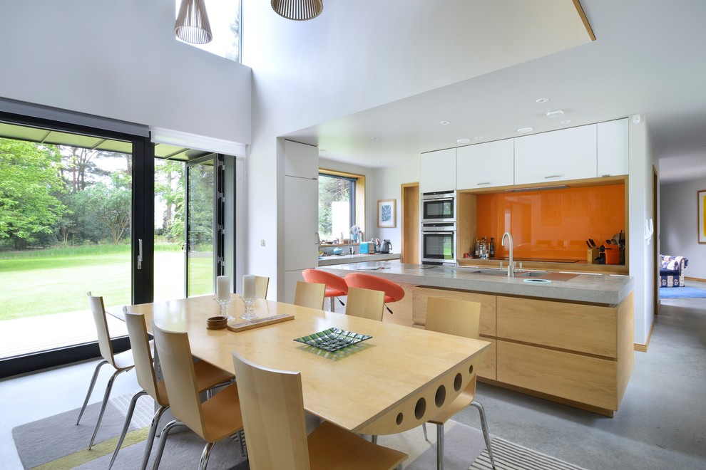 This is an example of a contemporary u-shaped open plan kitchen in Hampshire with orange splashback, with island, grey floor, grey benchtop, flat-panel cabinets, white cabinets, concrete benchtops, concrete floors, glass sheet splashback and stainless steel appliances.