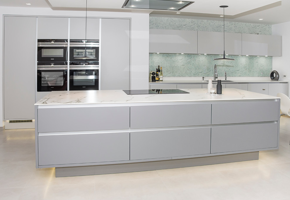 Large modern single-wall eat-in kitchen in Gloucestershire with an integrated sink, flat-panel cabinets, grey cabinets, solid surface benchtops, green splashback, glass sheet splashback, black appliances, porcelain floors and with island.