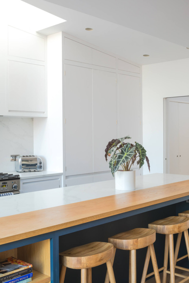 Photo of a large contemporary single-wall open plan kitchen in London with a drop-in sink, flat-panel cabinets, white cabinets, quartzite benchtops, white splashback, stone slab splashback, stainless steel appliances, ceramic floors, with island, white floor and white benchtop.