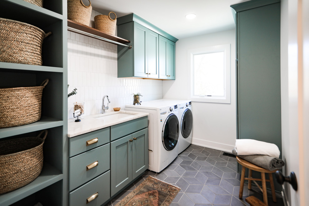 シカゴにある高級な中くらいなトランジショナルスタイルのおしゃれな洗濯室 (I型、アンダーカウンターシンク、シェーカースタイル扉のキャビネット、青いキャビネット、珪岩カウンター、白いキッチンパネル、サブウェイタイルのキッチンパネル、白い壁、セラミックタイルの床、左右配置の洗濯機・乾燥機、グレーの床、白いキッチンカウンター) の写真