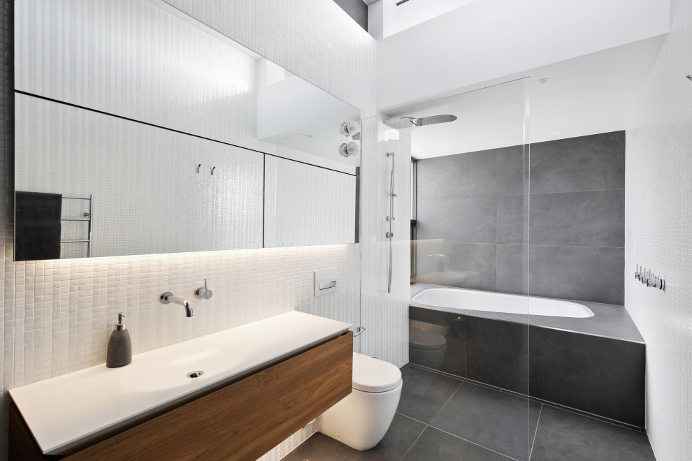 This is an example of a large contemporary master wet room bathroom in Sydney with flat-panel cabinets, medium wood cabinets, a drop-in tub, a wall-mount toilet, white tile, white walls, porcelain floors, an integrated sink, grey floor and white benchtops.