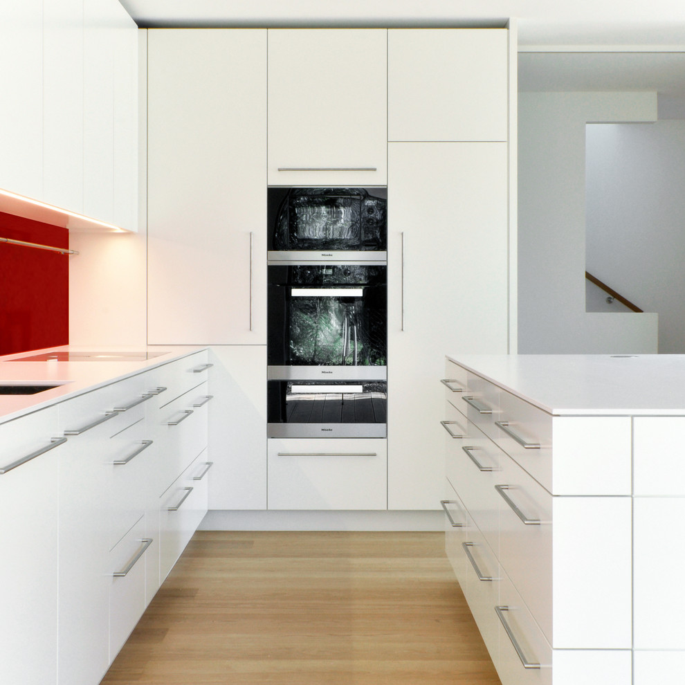 Mid-sized contemporary l-shaped open plan kitchen in Munich with flat-panel cabinets, white cabinets, red splashback, light hardwood floors, with island, an undermount sink and panelled appliances.