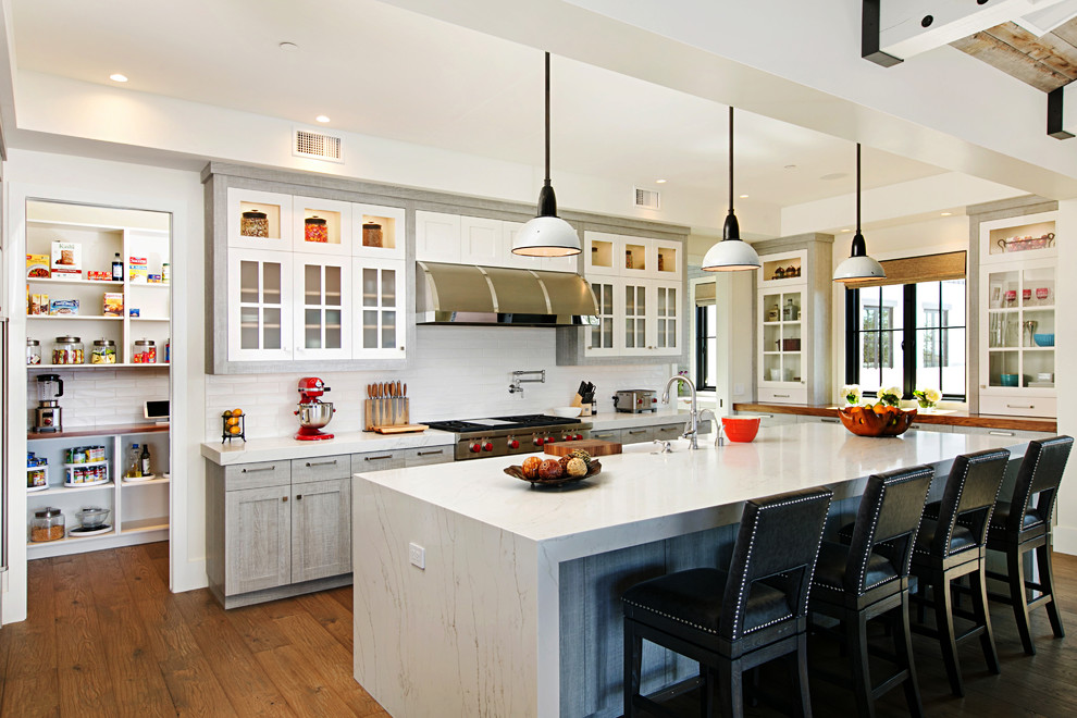 This is an example of a country l-shaped kitchen in San Diego with a farmhouse sink, glass-front cabinets, white cabinets, white splashback, stainless steel appliances, medium hardwood floors, with island, brown floor and white benchtop.