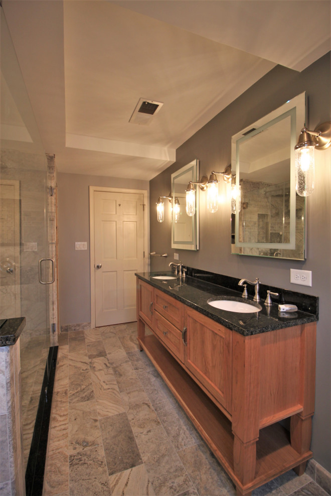 This is an example of a mid-sized transitional master bathroom in Indianapolis with flat-panel cabinets, medium wood cabinets, a double shower, a two-piece toilet, multi-coloured tile, stone tile, grey walls, travertine floors, an undermount sink, granite benchtops, a hinged shower door, black benchtops, a double vanity and a freestanding vanity.