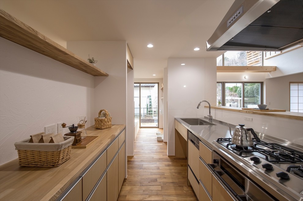 This is an example of an asian kitchen in Other with an integrated sink, flat-panel cabinets, light wood cabinets, stainless steel benchtops, white splashback, medium hardwood floors, a peninsula, brown floor and beige benchtop.
