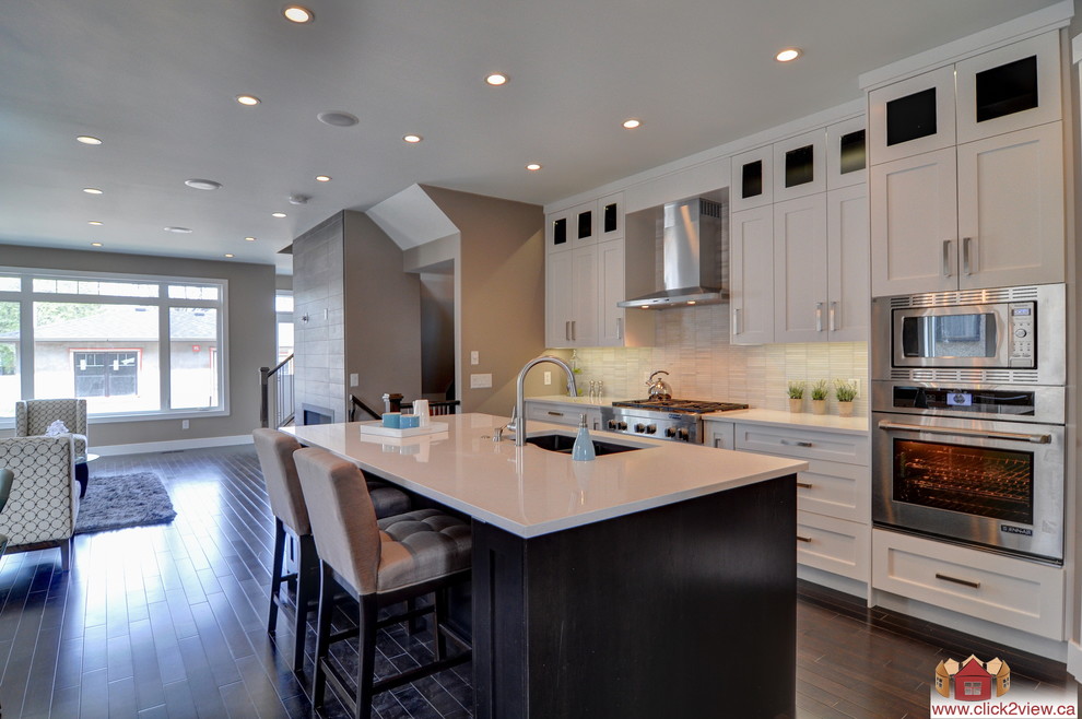 Photo of a mid-sized contemporary l-shaped eat-in kitchen in Calgary with a drop-in sink, recessed-panel cabinets, white cabinets, granite benchtops, beige splashback, ceramic splashback, stainless steel appliances, dark hardwood floors and with island.