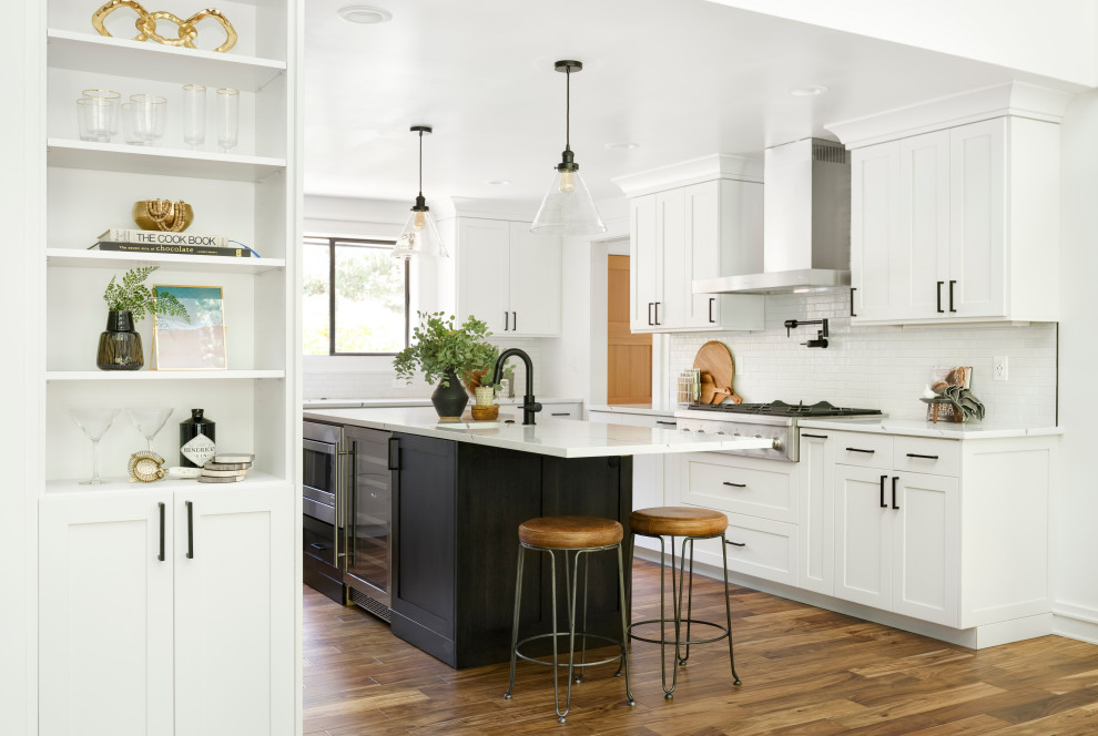 Mid-sized country u-shaped open plan kitchen in Portland with an undermount sink, shaker cabinets, white cabinets, quartz benchtops, white splashback, ceramic splashback, stainless steel appliances, medium hardwood floors, with island, brown floor and white benchtop.