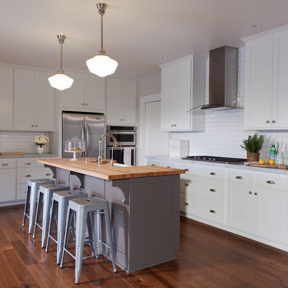 Photo of a country l-shaped kitchen in Santa Barbara with a farmhouse sink, shaker cabinets, white cabinets, white splashback, subway tile splashback, stainless steel appliances, dark hardwood floors and with island.