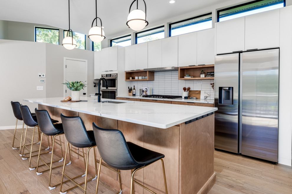 This is an example of an expansive midcentury galley open plan kitchen in Minneapolis with a single-bowl sink, flat-panel cabinets, white cabinets, quartz benchtops, white splashback, porcelain splashback, stainless steel appliances, light hardwood floors, with island, beige floor and white benchtop.