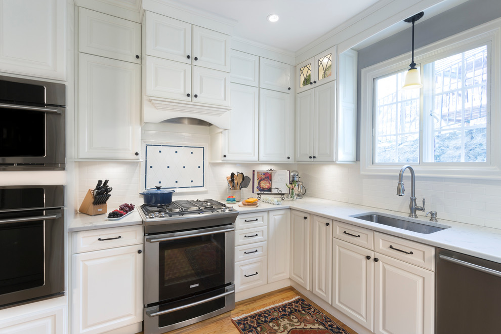 Mid-sized elegant l-shaped medium tone wood floor enclosed kitchen photo in New York with an undermount sink, raised-panel cabinets, white cabinets, quartz countertops, white backsplash, subway tile backsplash, stainless steel appliances and no island