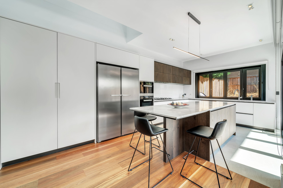 Design ideas for a large modern l-shaped eat-in kitchen in Sydney with a double-bowl sink, flat-panel cabinets, white cabinets, marble benchtops, white splashback, glass sheet splashback, stainless steel appliances, medium hardwood floors, with island, brown floor and grey benchtop.