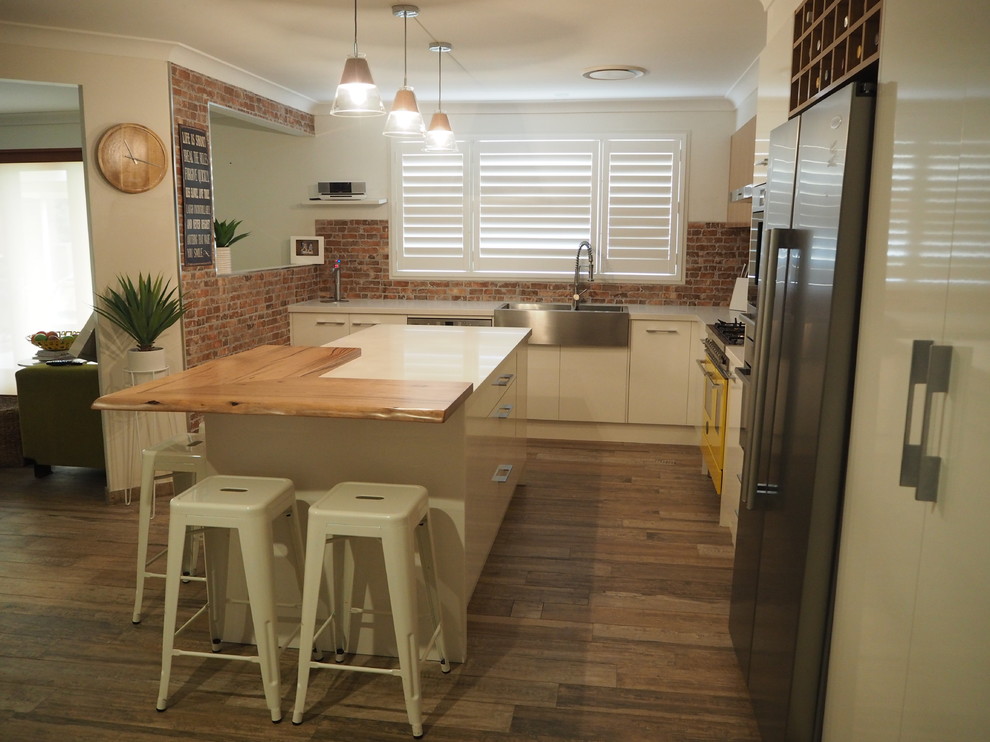 Inspiration for a mid-sized eclectic l-shaped eat-in kitchen in Sydney with flat-panel cabinets, white cabinets, quartz benchtops, with island, a farmhouse sink, red splashback, brick splashback, stainless steel appliances, medium hardwood floors and white benchtop.