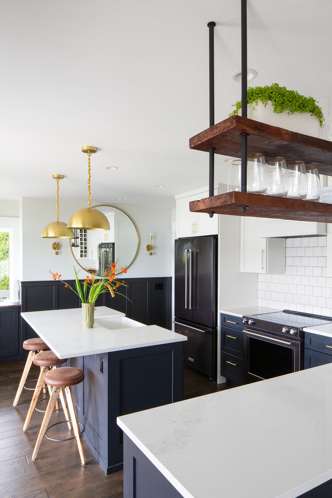 Inspiration for an eclectic l-shaped kitchen in Seattle with a farmhouse sink, shaker cabinets, blue cabinets, white splashback, subway tile splashback, black appliances, dark hardwood floors, with island and white benchtop.