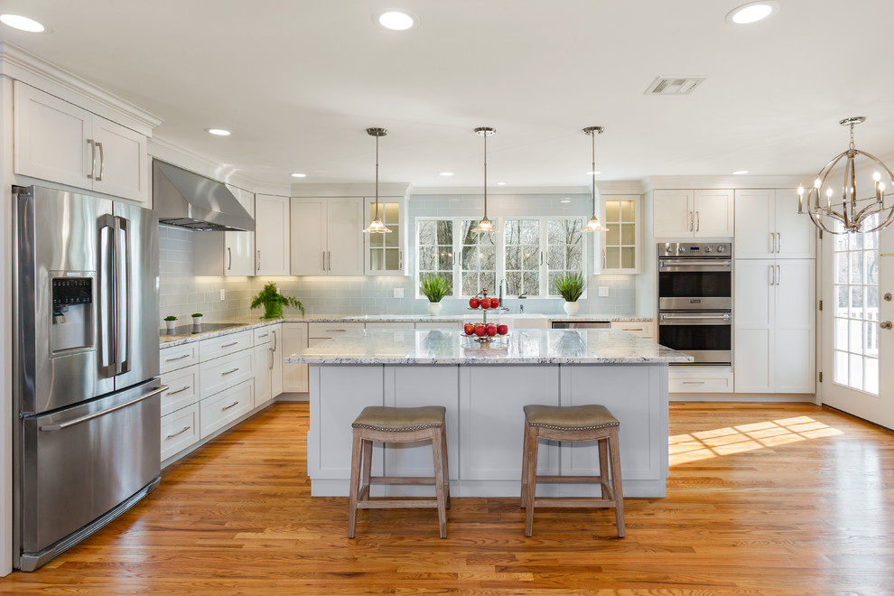 This is an example of a large transitional l-shaped eat-in kitchen in New York with a farmhouse sink, shaker cabinets, white cabinets, granite benchtops, blue splashback, glass tile splashback, stainless steel appliances, medium hardwood floors, with island, brown floor and grey benchtop.