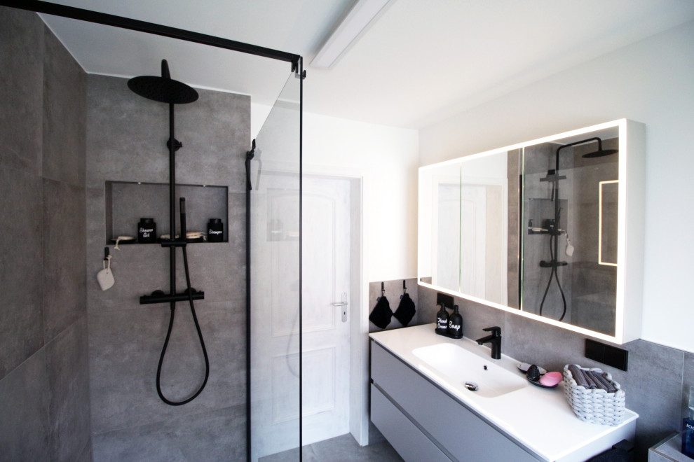 Inredning av ett modernt badrum, med grå skåp, en kantlös dusch, en toalettstol med separat cisternkåpa, grå kakel, vita väggar och grått golv
