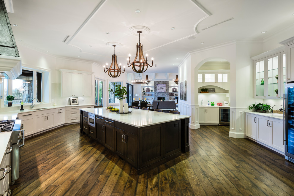Huge mountain style u-shaped medium tone wood floor and brown floor open concept kitchen photo in Vancouver with an undermount sink, shaker cabinets, white cabinets, solid surface countertops, white backsplash, stone slab backsplash, stainless steel appliances, an island and white countertops
