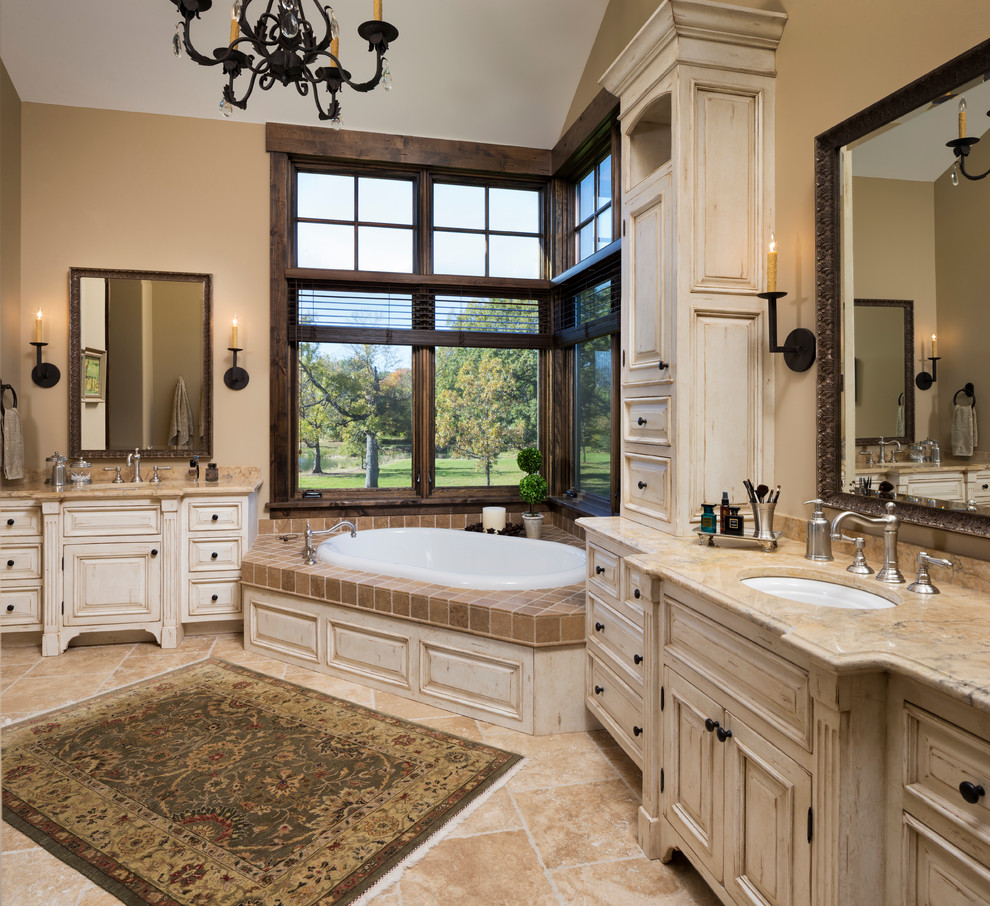 Inspiration for a large country master bathroom in Minneapolis with an undermount sink, raised-panel cabinets, distressed cabinets, marble benchtops, a drop-in tub, beige tile, stone tile, beige walls, travertine floors and beige floor.