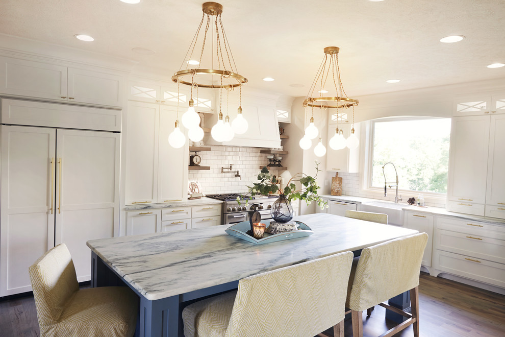 Example of a huge mountain style l-shaped medium tone wood floor eat-in kitchen design with a farmhouse sink, shaker cabinets, white cabinets, white backsplash, subway tile backsplash, paneled appliances, an island and white countertops