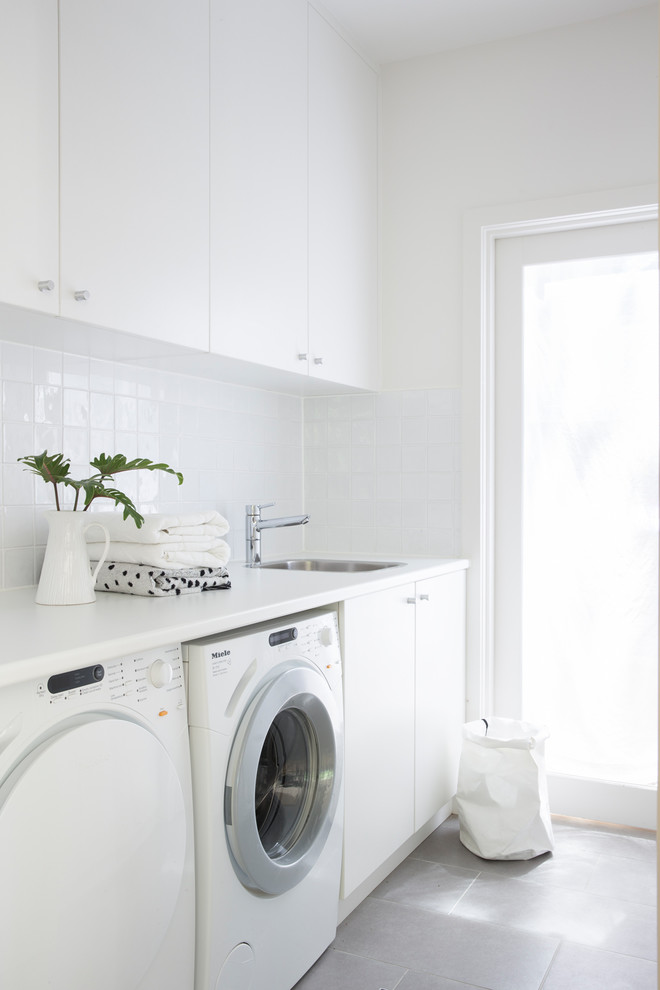 This is an example of a contemporary single-wall dedicated laundry room in Sydney with a drop-in sink, flat-panel cabinets, white cabinets, white walls, a side-by-side washer and dryer, grey floor and white benchtop.