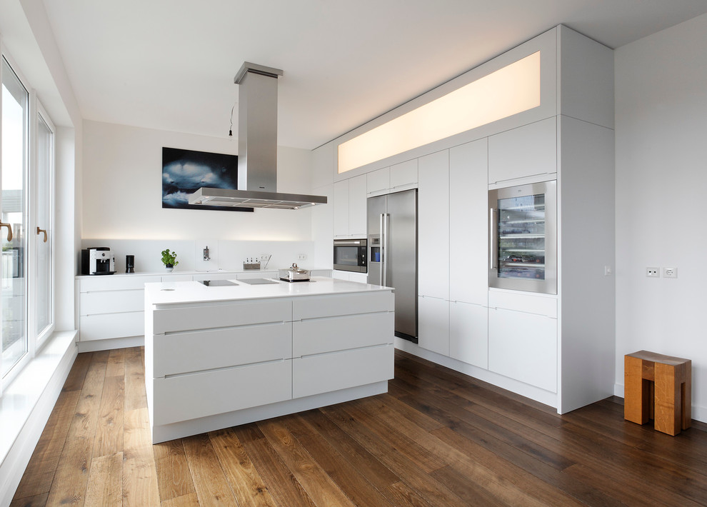 Design ideas for a large contemporary l-shaped open plan kitchen in Hamburg with flat-panel cabinets, white cabinets, white splashback, stainless steel appliances, dark hardwood floors, with island, solid surface benchtops, a drop-in sink, glass sheet splashback and brown floor.