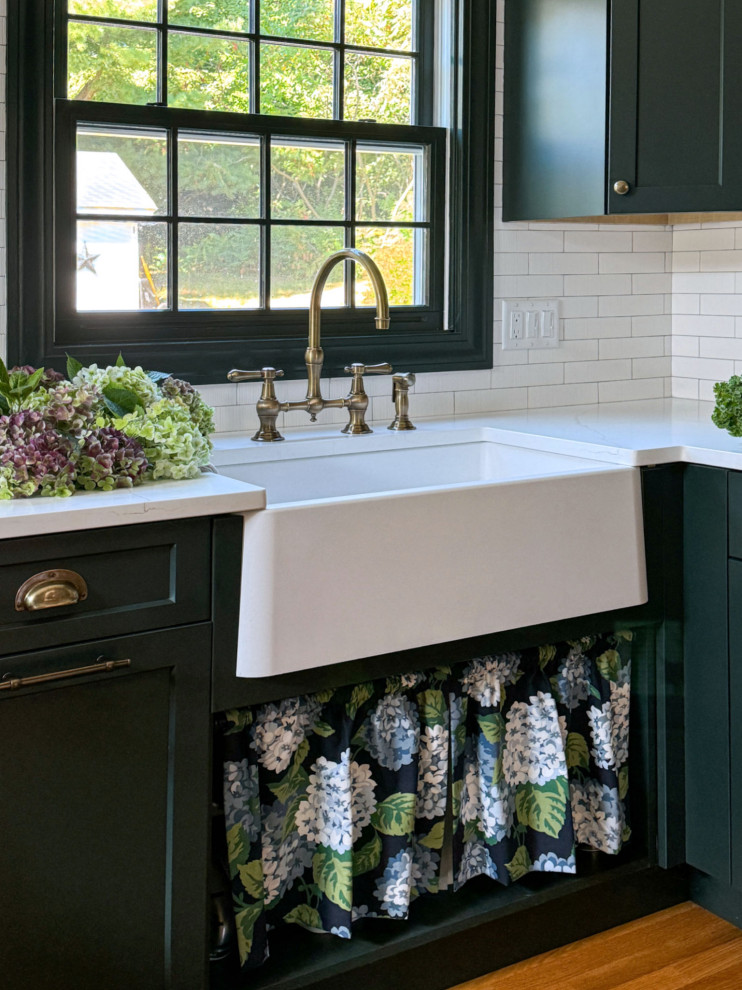 Example of a transitional u-shaped medium tone wood floor and brown floor kitchen design in Boston with a farmhouse sink, shaker cabinets, green cabinets, quartz countertops, white backsplash, ceramic backsplash, black appliances, a peninsula and white countertops