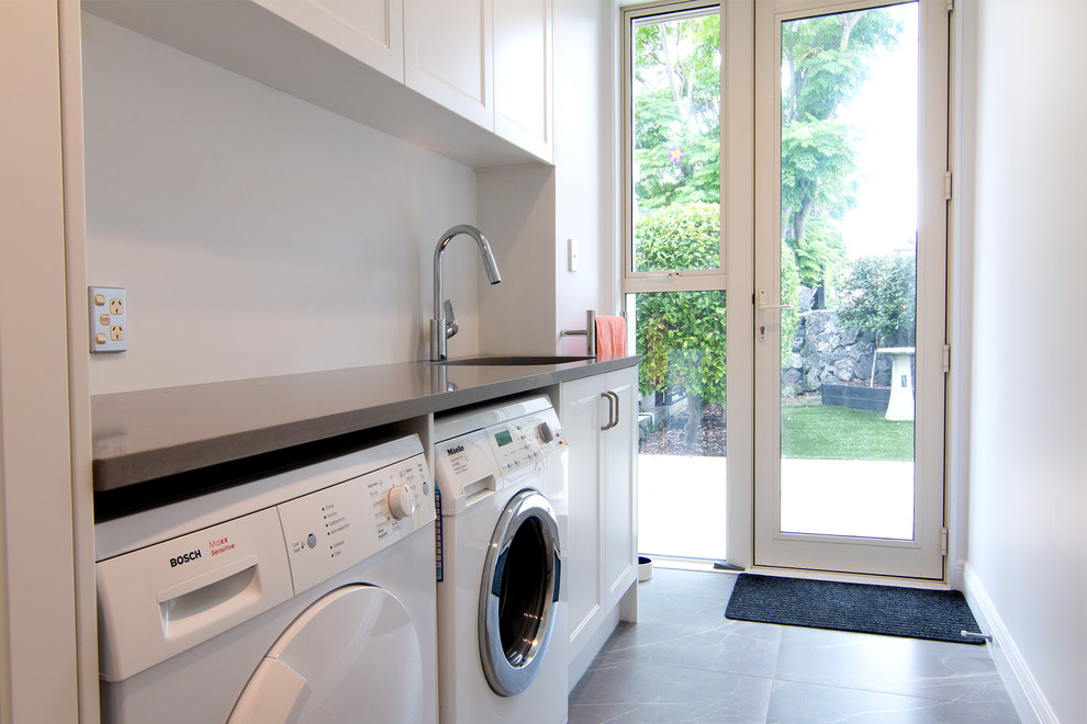 Inspiration for a mid-sized contemporary galley dedicated laundry room in Auckland with an undermount sink, quartz benchtops, white splashback, white walls, porcelain floors, a side-by-side washer and dryer, grey floor and grey benchtop.