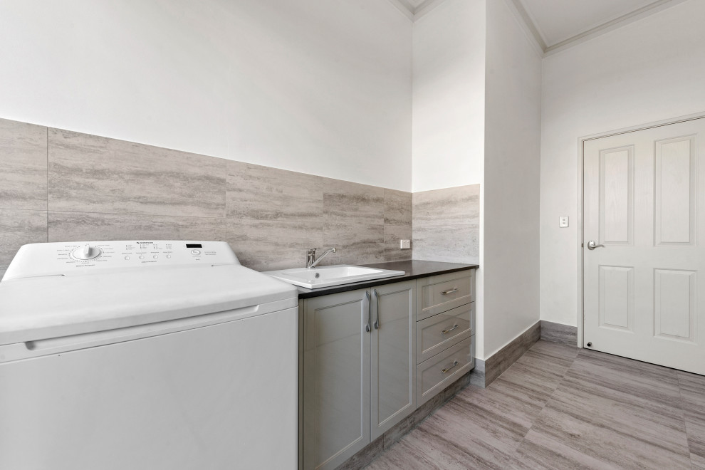 Design ideas for a mid-sized traditional single-wall dedicated laundry room in Perth with a drop-in sink, recessed-panel cabinets, grey cabinets, white walls, grey floor and black benchtop.