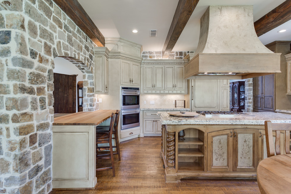 Mid-sized traditional l-shaped eat-in kitchen in Dallas with an undermount sink, raised-panel cabinets, distressed cabinets, granite benchtops, white splashback, travertine splashback, stainless steel appliances, medium hardwood floors, with island and brown floor.