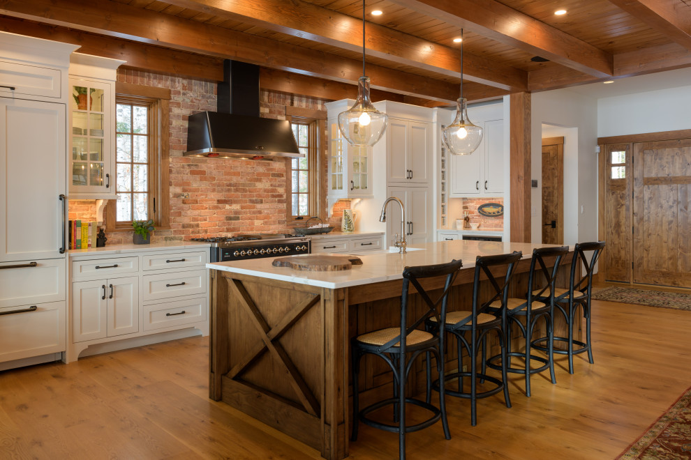 This is an example of a large country u-shaped kitchen in Other with a farmhouse sink, shaker cabinets, white cabinets, red splashback, brick splashback, panelled appliances, medium hardwood floors, with island, brown floor, white benchtop and wood.