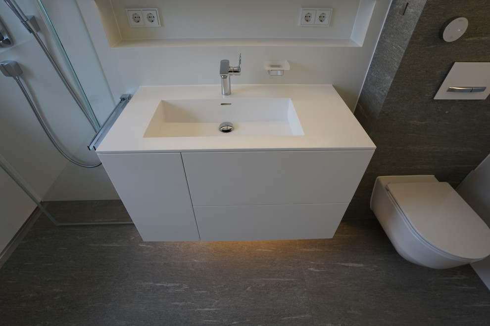 Duschbad With flächenbündigen Schrankfronten, weißen Schränken, bodengleicher Dusche, Wandtoilette, weißen Fliesen, Steinfliesen, Wandwaschbecken, Mineralwerkstoff-Waschtisch, grauem Boden, Falttür-Duschabtrennung, weißer Waschtischplatte, Einzelwaschbecken and schwebendem Waschtisch in Munich