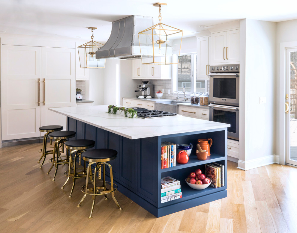 Photo of a mid-sized transitional l-shaped open plan kitchen in New York with a farmhouse sink, shaker cabinets, white cabinets, quartz benchtops, grey splashback, subway tile splashback, panelled appliances, light hardwood floors, with island, brown floor and white benchtop.