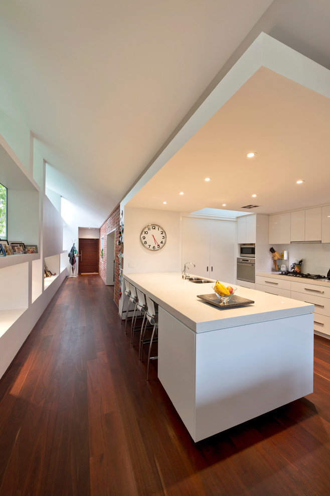Inspiration for a mid-sized contemporary galley eat-in kitchen in Perth with an undermount sink, flat-panel cabinets, white cabinets, solid surface benchtops, white splashback, medium hardwood floors, with island, glass sheet splashback and stainless steel appliances.