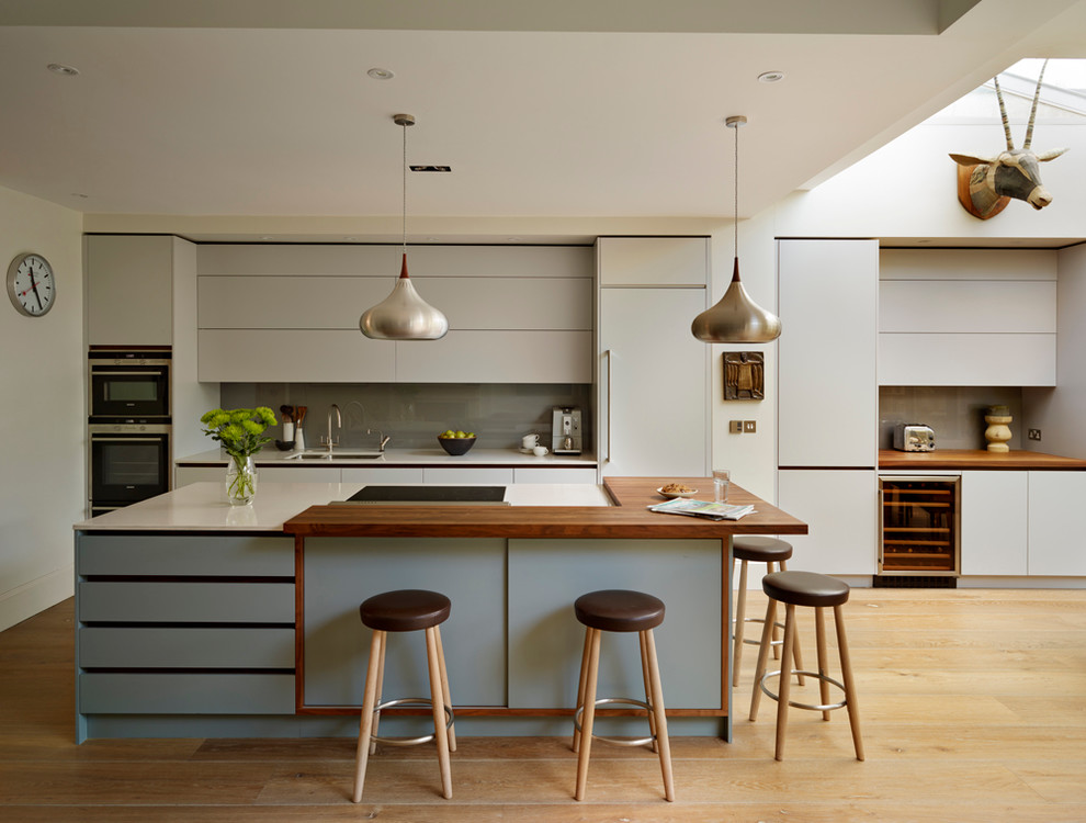 Large contemporary galley open plan kitchen in London with an undermount sink, flat-panel cabinets, solid surface benchtops, glass sheet splashback, medium hardwood floors, with island, grey cabinets, grey splashback and panelled appliances.