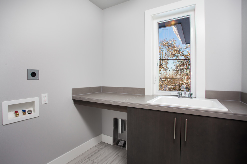 This is an example of a mid-sized modern single-wall utility room in Portland with a drop-in sink, flat-panel cabinets, dark wood cabinets, tile benchtops, grey walls, ceramic floors, a side-by-side washer and dryer and grey floor.