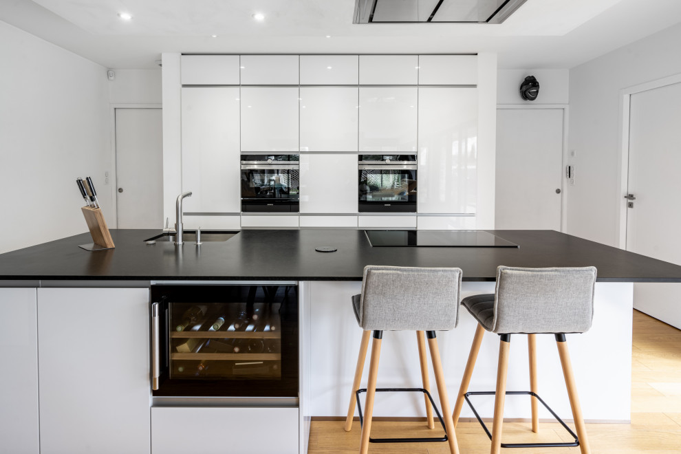 Small contemporary single-wall open plan kitchen in Nantes with an undermount sink, flat-panel cabinets, white cabinets, granite benchtops, panelled appliances, light hardwood floors, with island, brown floor and black benchtop.