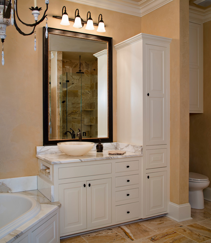 Photo of a large traditional master bathroom in New Orleans with a vessel sink, beaded inset cabinets, white cabinets, marble benchtops, a corner tub, a corner shower, a one-piece toilet, beige tile, stone tile, beige walls and marble floors.