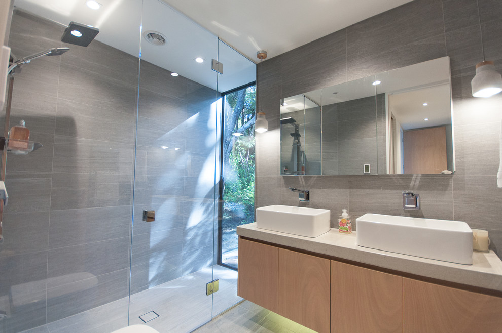This is an example of a mid-sized contemporary bathroom in Auckland with light wood cabinets, an open shower, a wall-mount toilet, gray tile, stone tile, grey walls, ceramic floors and concrete benchtops.