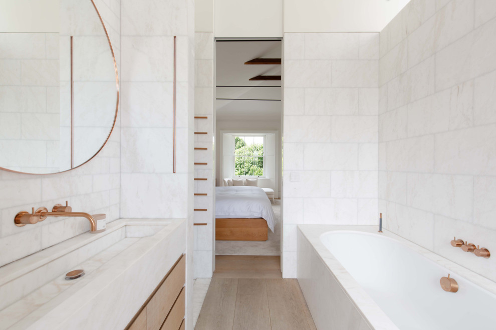 This is an example of a contemporary bathroom in London with flat-panel cabinets, medium wood cabinets, an undermount tub, white tile, white walls, light hardwood floors, a trough sink, beige floor, white benchtops and a single vanity.