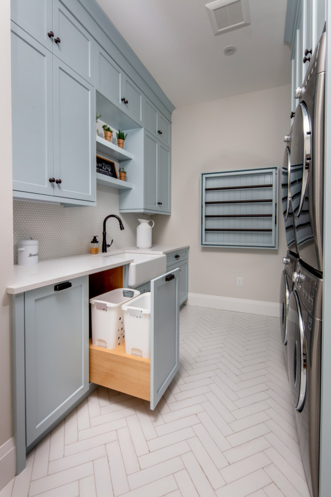 Design ideas for a mid-sized country galley dedicated laundry room in Toronto with a farmhouse sink, shaker cabinets, blue cabinets, quartz benchtops, grey splashback, ceramic splashback, beige walls, a stacked washer and dryer, white floor and white benchtop.