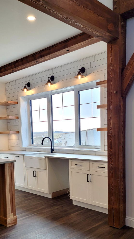 Photo of a large farmhouse single-wall kitchen/diner in Edmonton with a belfast sink, beaded cabinets, white cabinets, engineered stone countertops, white splashback, metro tiled splashback, vinyl flooring, an island, brown floors, grey worktops and exposed beams.
