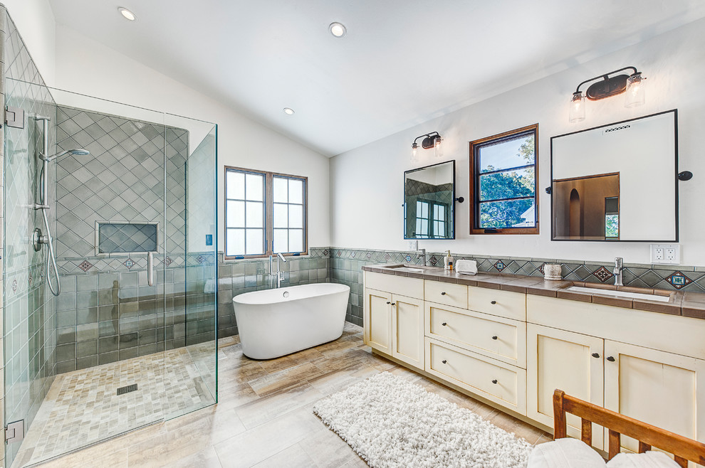 Large mediterranean master bathroom in Santa Barbara with shaker cabinets, distressed cabinets, a freestanding tub, a corner shower, a two-piece toilet, gray tile, ceramic tile, white walls, travertine floors, tile benchtops, beige floor and a hinged shower door.