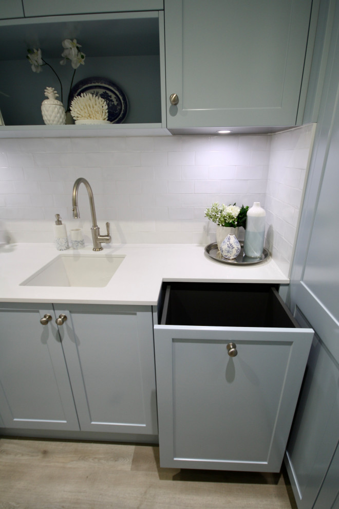 This is an example of a mid-sized transitional u-shaped dedicated laundry room in Sydney with an undermount sink, shaker cabinets, blue cabinets, quartz benchtops, white splashback, subway tile splashback, white walls, light hardwood floors, a stacked washer and dryer, brown floor and white benchtop.