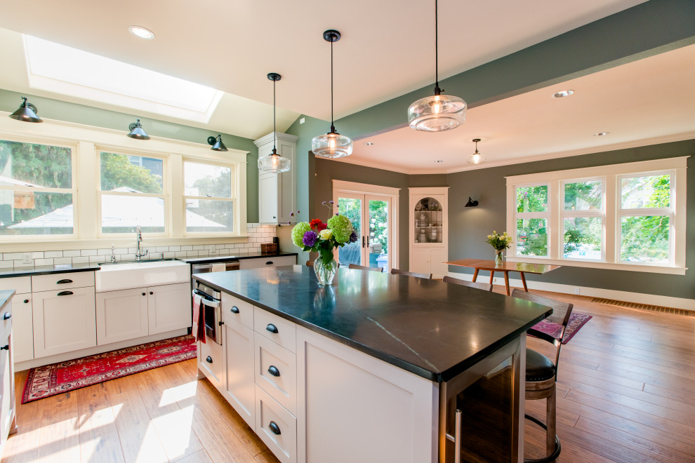 Photo of a large arts and crafts u-shaped eat-in kitchen in Seattle with a farmhouse sink, shaker cabinets, white cabinets, quartz benchtops, white splashback, brick splashback, stainless steel appliances, medium hardwood floors, with island, brown floor, black benchtop and vaulted.