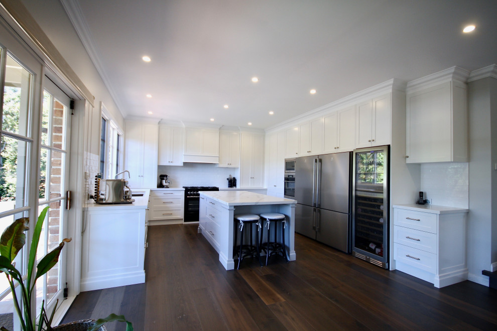 This is an example of a large transitional u-shaped open plan kitchen in Sydney with a drop-in sink, shaker cabinets, white cabinets, quartz benchtops, yellow splashback, subway tile splashback, black appliances, dark hardwood floors, with island, brown floor and white benchtop.