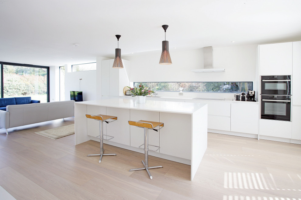 This is an example of a modern galley kitchen in Surrey with flat-panel cabinets, white cabinets, black appliances, light hardwood floors, with island, beige floor and white benchtop.